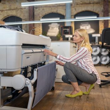 At workshop. Blonde woman crouching at printing machine, checking something